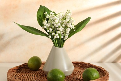 Photo of Beautiful lilies of the valley flowers in vase and limes on white table near beige wall
