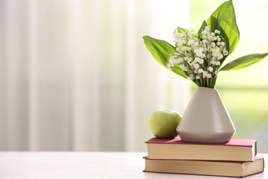 Photo of Beautiful lilies of the valley flowers in vase, apple and books on white table against blurred background. Space for text