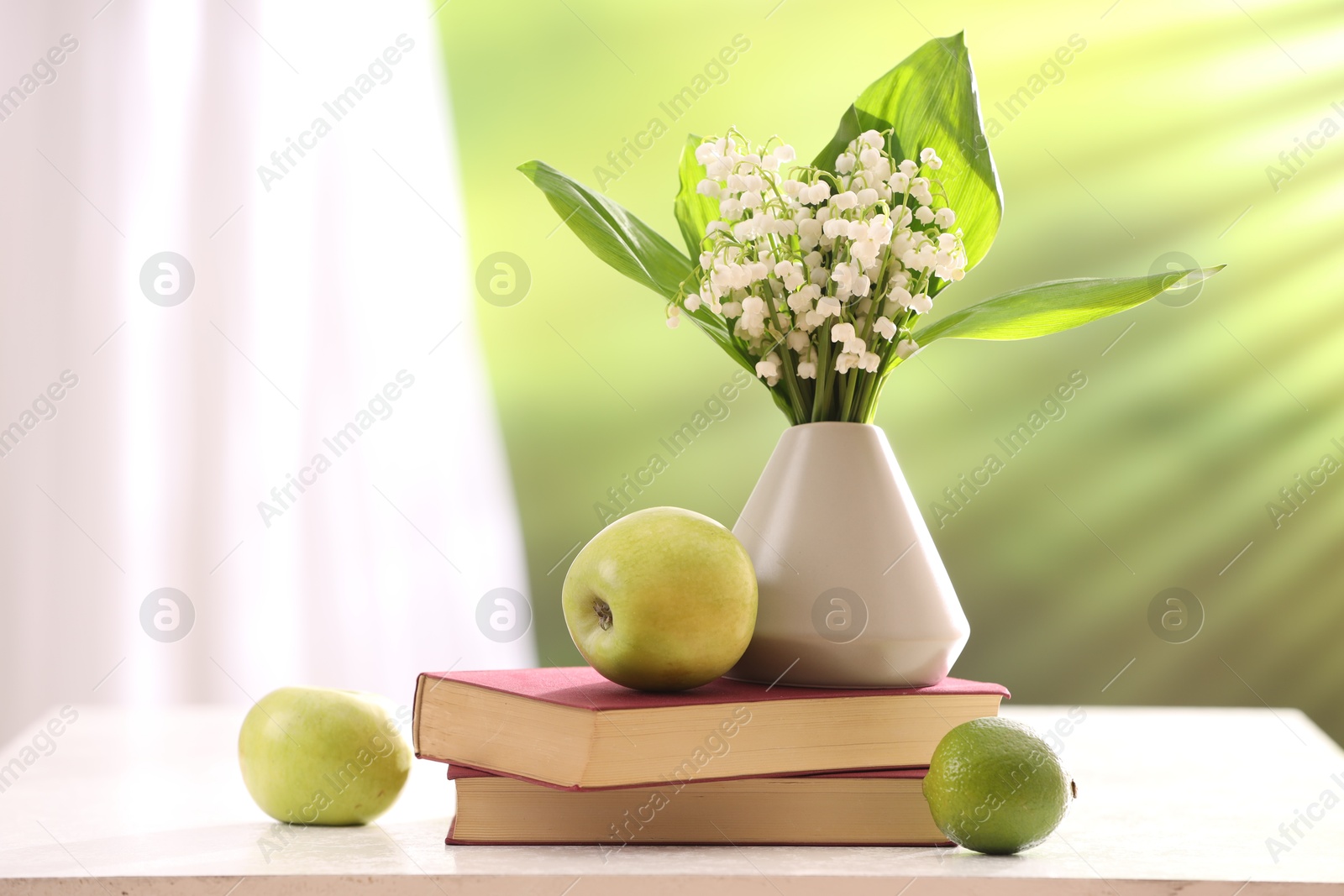 Beautiful lilies of the valley flowers in vase, fruits and books on white table against blurred background. Space for text Photo of Beautiful lilies of the valley flowers in vase, fruits and books on white table against blurred background. Space for text