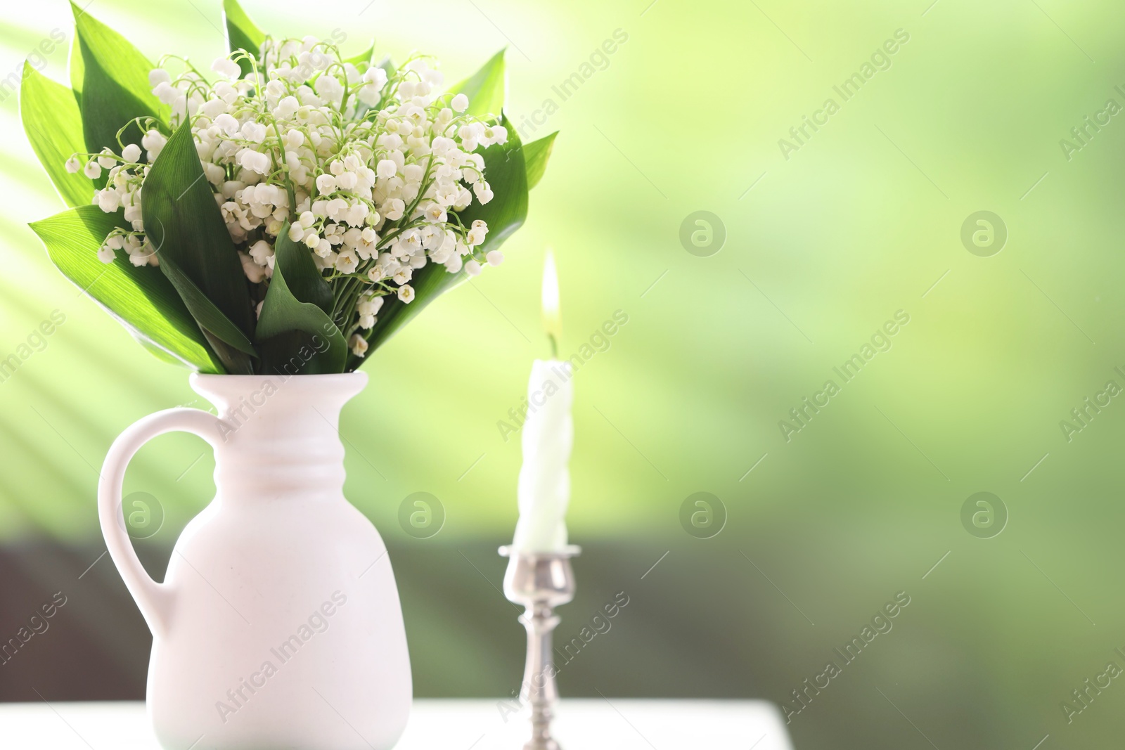 Beautiful lilies of the valley flowers in vase and candle on white table against blurred background. Space for text Photo of Beautiful lilies of the valley flowers in vase and candle on white table against blurred background. Space for text