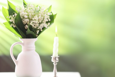 Photo of Beautiful lilies of the valley flowers in vase and candle on white table against blurred background. Space for text