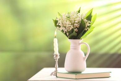 Photo of Beautiful lilies of the valley flowers in vase, candle and book on white table against blurred background. Space for text