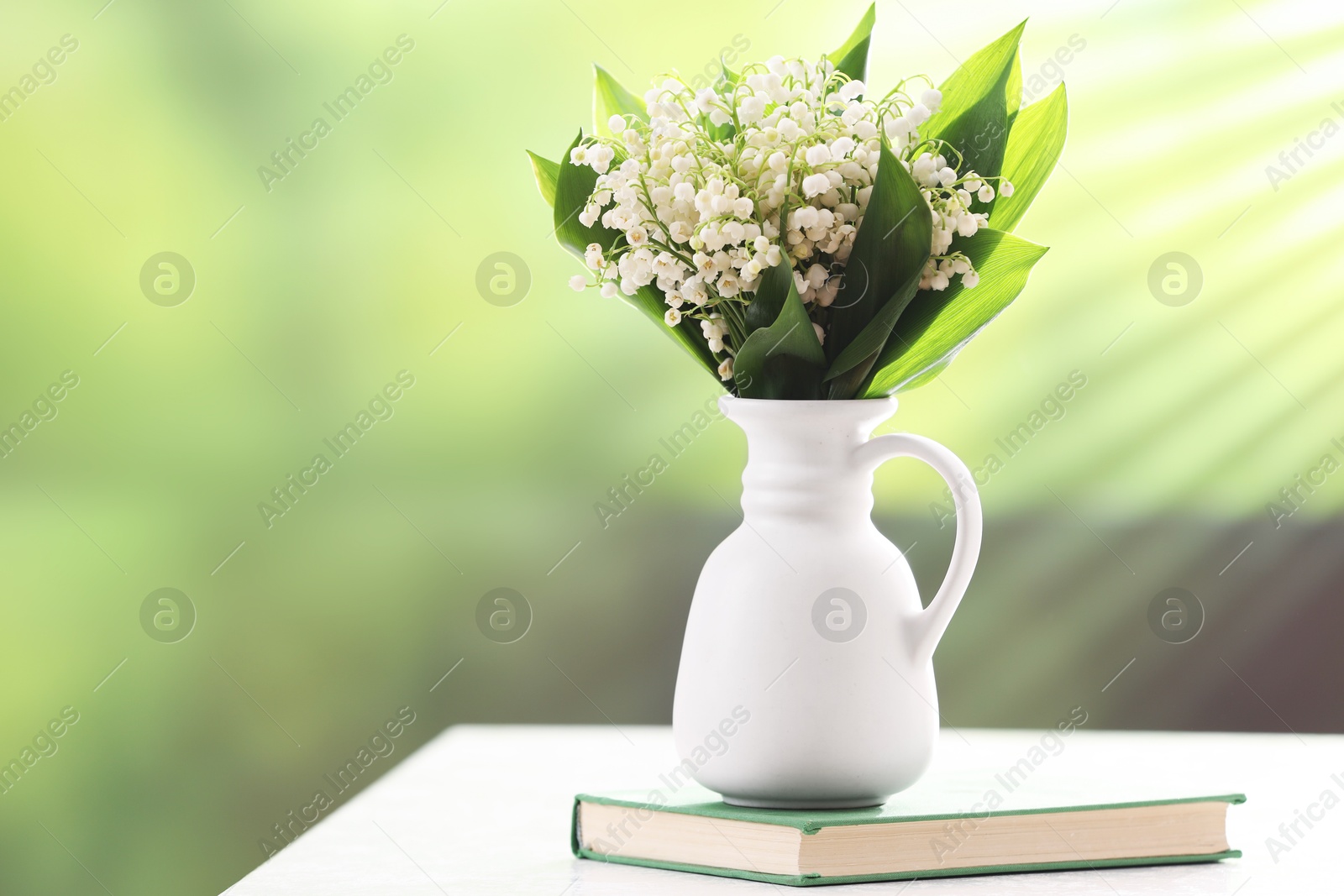 Beautiful lilies of the valley flowers in vase and book on white table against blurred background. Space for text Photo of Beautiful lilies of the valley flowers in vase and book on white table against blurred background. Space for text