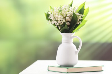 Photo of Beautiful lilies of the valley flowers in vase and book on white table against blurred background. Space for text