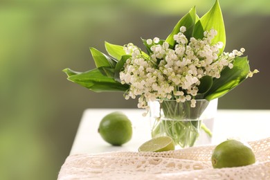 Photo of Beautiful lilies of the valley flowers in vase and limes on white table against blurred background