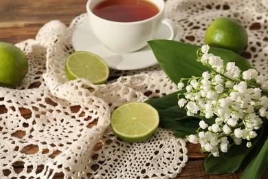 Photo of Beautiful lilies of the valley flowers, tea and limes on wooden table, closeup