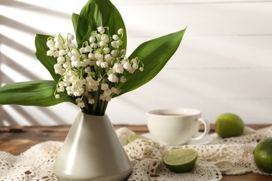 Photo of Beautiful lilies of the valley flowers in vase, tea and limes on wooden table, closeup