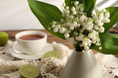 Photo of Beautiful lilies of the valley flowers in vase, tea and limes on wooden table, closeup