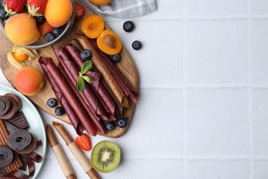 Photo of Tasty candied leather rolls, fruits and berries on white tiled table, flat lay. Space for text