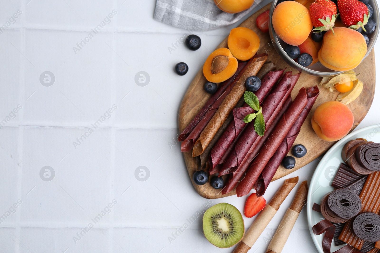 Tasty candied leather rolls, fruits and berries on white tiled table, flat lay. Space for text Photo of Tasty candied leather rolls, fruits and berries on white tiled table, flat lay. Space for text