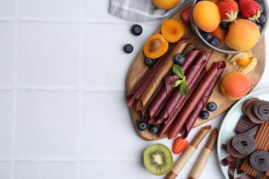 Photo of Tasty candied leather rolls, fruits and berries on white tiled table, flat lay. Space for text