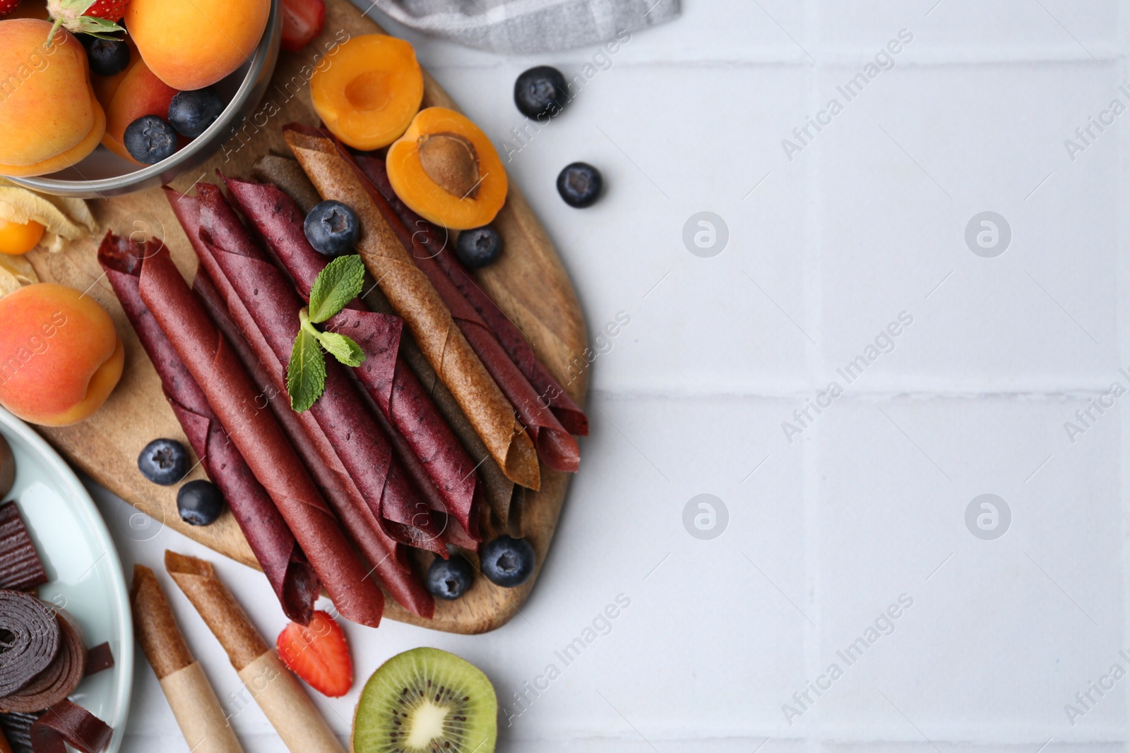 Tasty candied leather rolls, fruits and berries on white tiled table, flat lay. Space for text Photo of Tasty candied leather rolls, fruits and berries on white tiled table, flat lay. Space for text