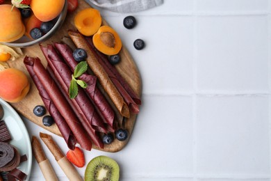Photo of Tasty candied leather rolls, fruits and berries on white tiled table, flat lay. Space for text