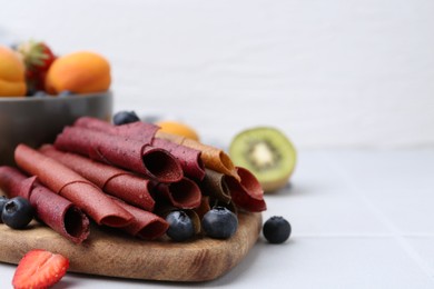 Photo of Tasty candied leather rolls, fruits and berries on white tiled table, closeup. Space for text