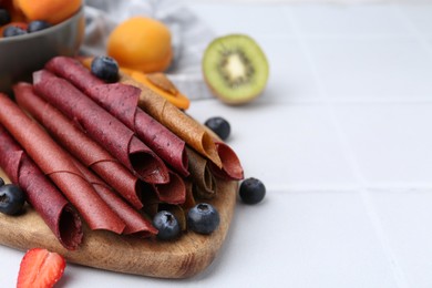 Photo of Tasty candied leather rolls, fruits and berries on white tiled table, closeup. Space for text
