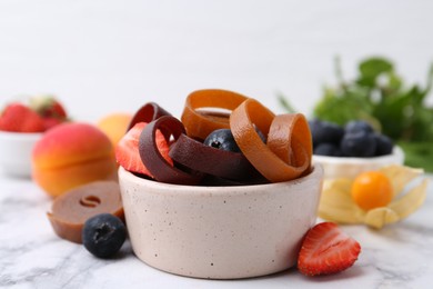 Photo of Tasty fruit leather rolls in bowl and berries on white marble table, closeup