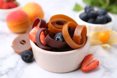 Photo of Tasty fruit leather rolls in bowl and berries on white marble table, closeup