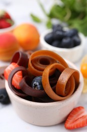 Photo of Tasty fruit leather rolls in bowl and berries on white marble table, closeup
