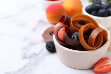 Photo of Tasty fruit leather rolls in bowl and berries on white marble table, closeup. Space for text