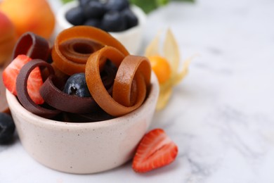 Photo of Tasty fruit leather rolls in bowl and berries on white marble table, closeup. Space for text