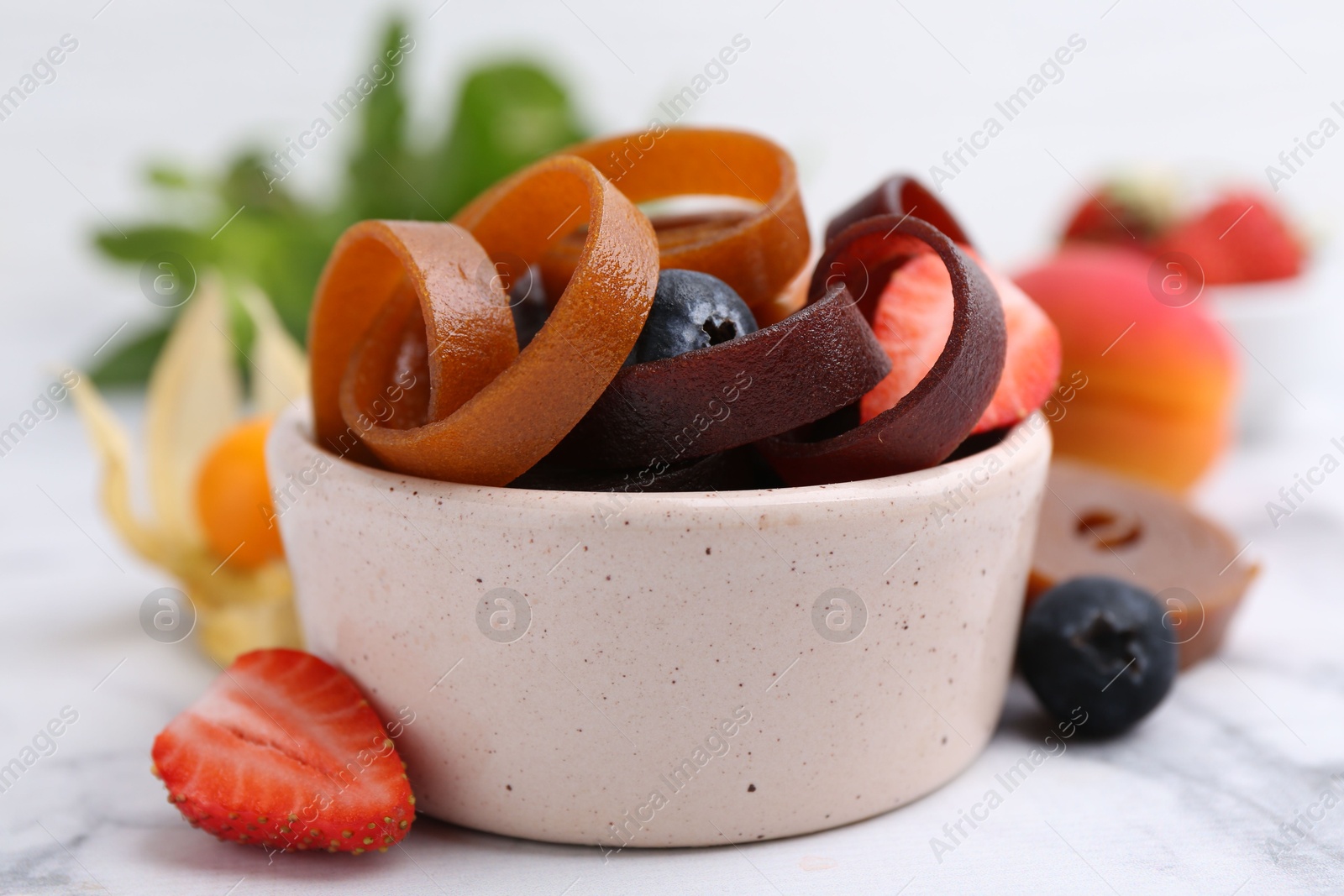 Tasty fruit leather rolls in bowl and berries on white marble table, closeup Photo of Tasty fruit leather rolls in bowl and berries on white marble table, closeup