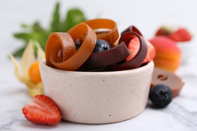 Photo of Tasty fruit leather rolls in bowl and berries on white marble table, closeup