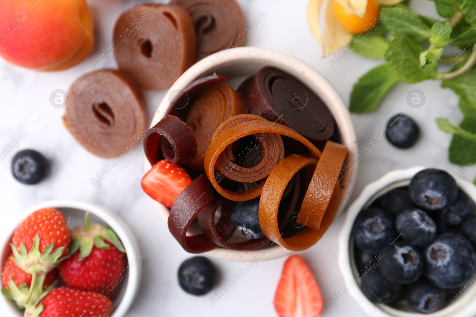 Tasty fruit leather rolls in bowl, mint and berries on white marble table, flat lay Photo of Tasty fruit leather rolls in bowl, mint and berries on white marble table, flat lay