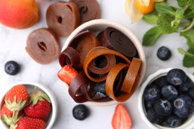 Photo of Tasty fruit leather rolls in bowl, mint and berries on white marble table, flat lay