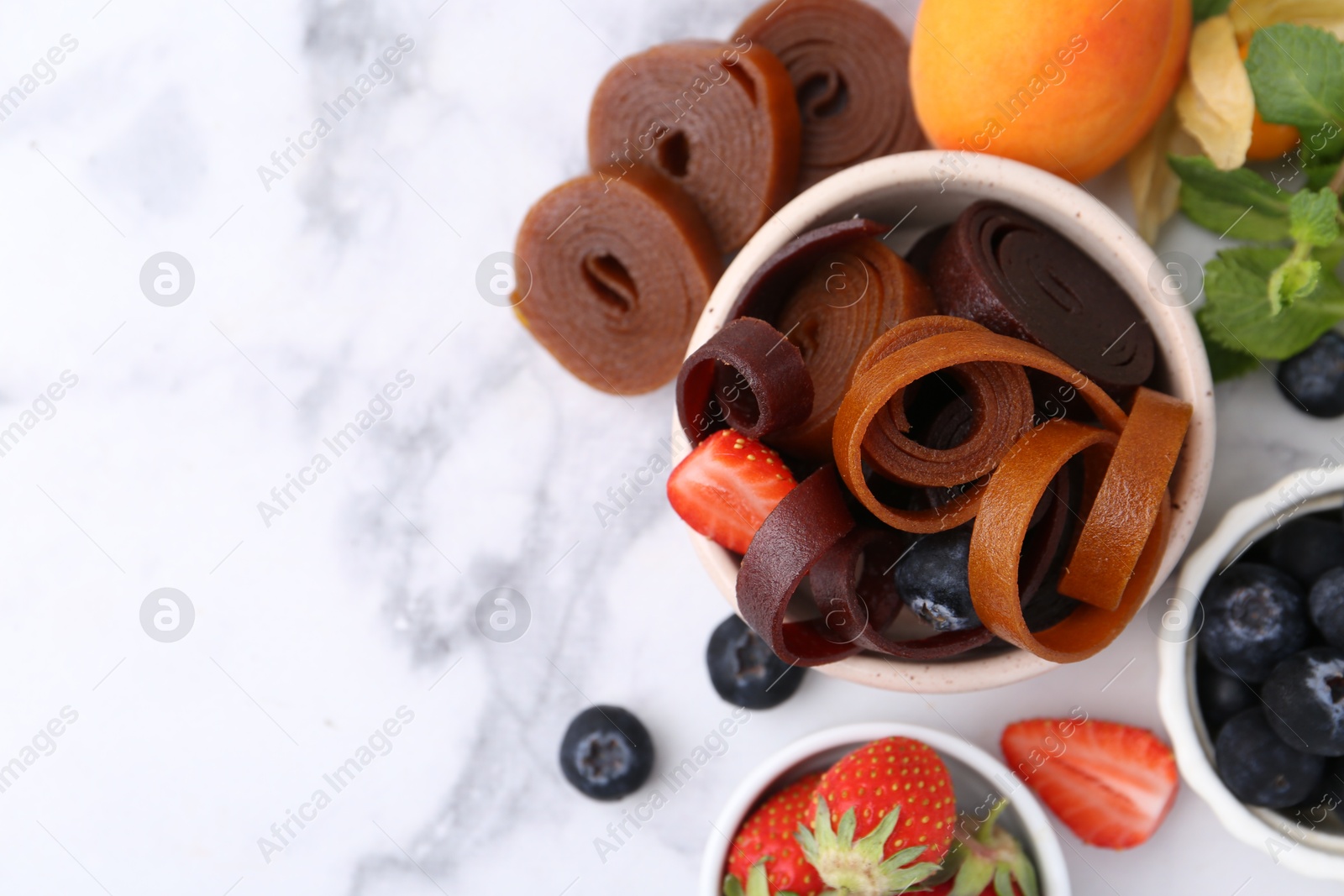 Tasty fruit leather rolls in bowl, mint and berries on white marble table, flat lay. Space for text Photo of Tasty fruit leather rolls in bowl, mint and berries on white marble table, flat lay. Space for text