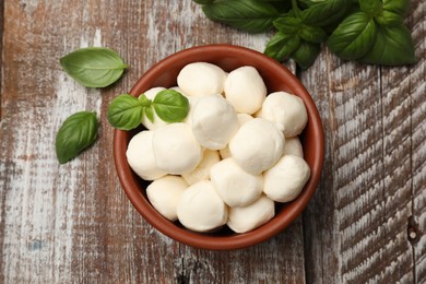 Photo of Tasty mozzarella cheese balls in bowl and basil on wooden table, flat lay