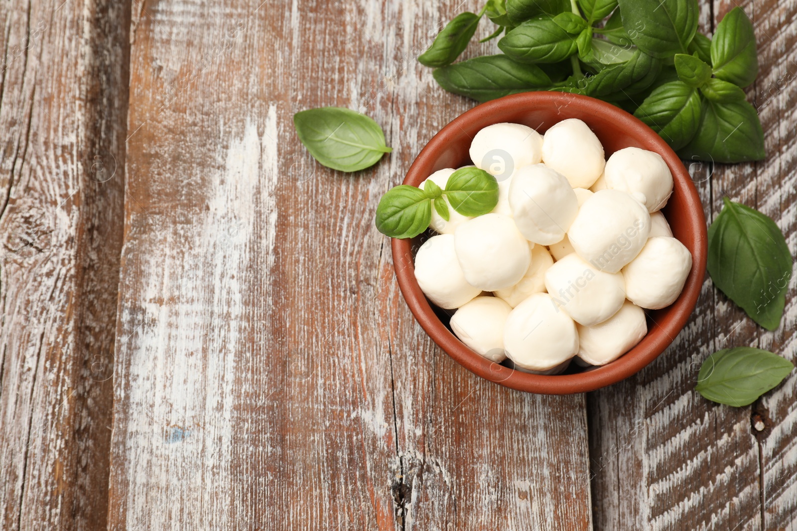 Photo of Tasty mozzarella cheese balls in bowl and basil on wooden table, flat lay. Space for text