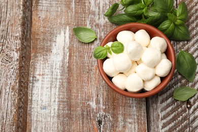 Photo of Tasty mozzarella cheese balls in bowl and basil on wooden table, flat lay. Space for text