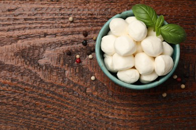 Photo of Tasty mozzarella cheese balls in bowl, peppercorns and basil on wooden table, top view. Space for text