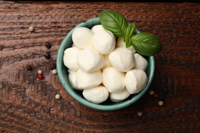 Photo of Tasty mozzarella cheese balls in bowl, peppercorns and basil on wooden table, top view