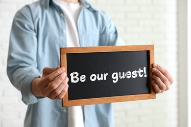 Image of Man holding small blackboard with phrase Be Our Guest indoors, closeup