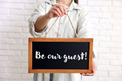 Image of Woman holding small blackboard with phrase Be Our Guest indoors, closeup