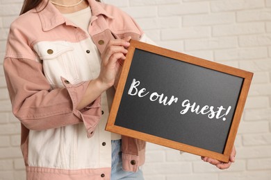 Image of Woman holding small blackboard with phrase Be Our Guest indoors, closeup