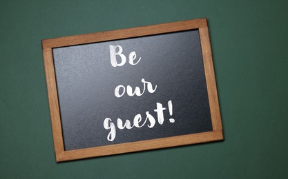 Image of Small blackboard with phrase Be Our Guest on dark green background, top view