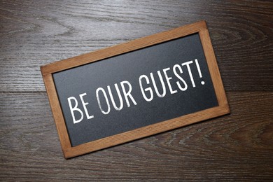 Image of Small blackboard with phrase Be Our Guest on wooden background, top view