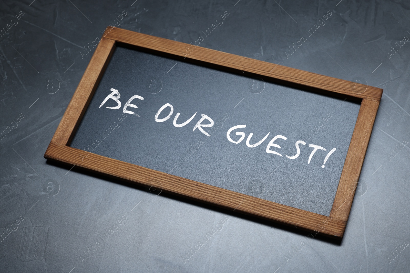 Small blackboard with phrase Be Our Guest on dark gray textured background, above view Image of Small blackboard with phrase Be Our Guest on dark gray textured background, above view