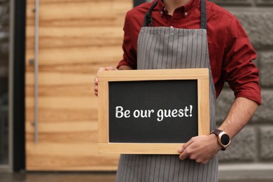 Image of Man holding small blackboard with phrase Be Our Guest outdoors, closeup