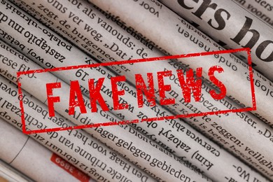 Image of Red Fake News stamp and newspapers in different languages as background, closeup