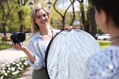 Photo of Professional photographer with reflector and camera near model during shooting outdoors on sunny day