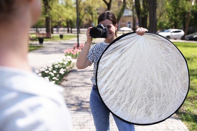 Photo of Professional photographer with reflector and camera near model during shooting outdoors on sunny day