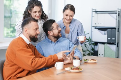 Photo of Man showing his colleagues something on smartphone during lunch break in office