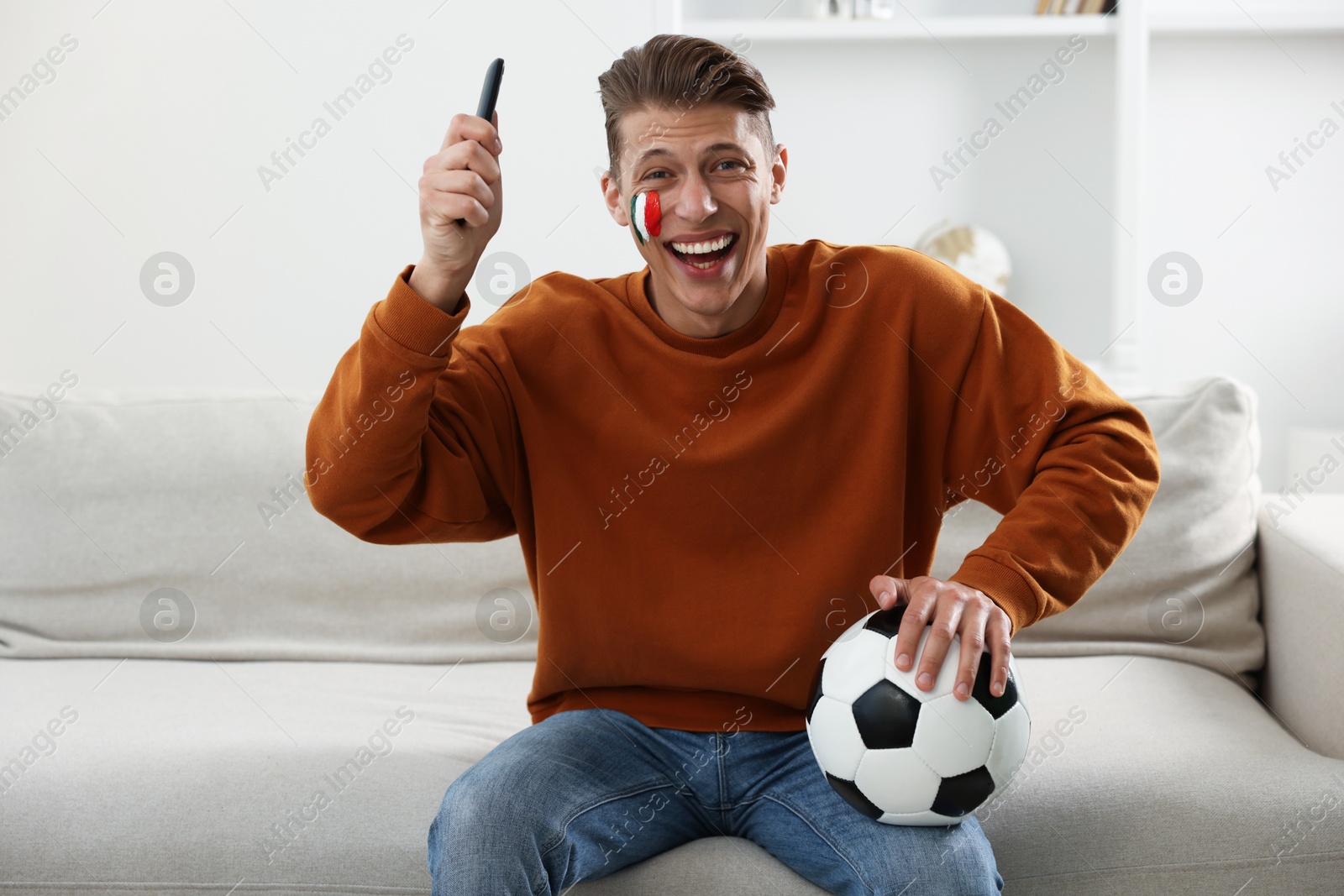 Football fan with remote control and soccer ball watching game on sofa indoors Photo of Football fan with remote control and soccer ball watching game on sofa indoors