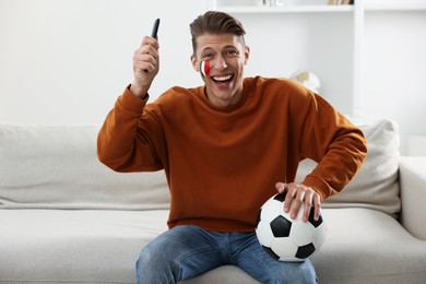 Photo of Football fan with remote control and soccer ball watching game on sofa indoors