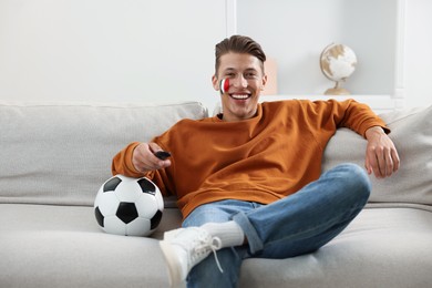 Photo of Football fan with remote control and soccer ball watching game on sofa indoors