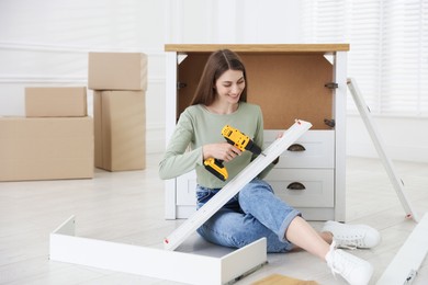 Photo of Smiling woman assembling furniture with electric screwdriver on floor at home
