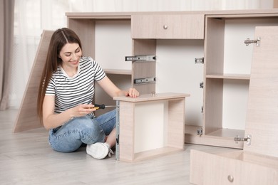 Photo of Smiling woman assembling furniture with screwdriver on floor at home